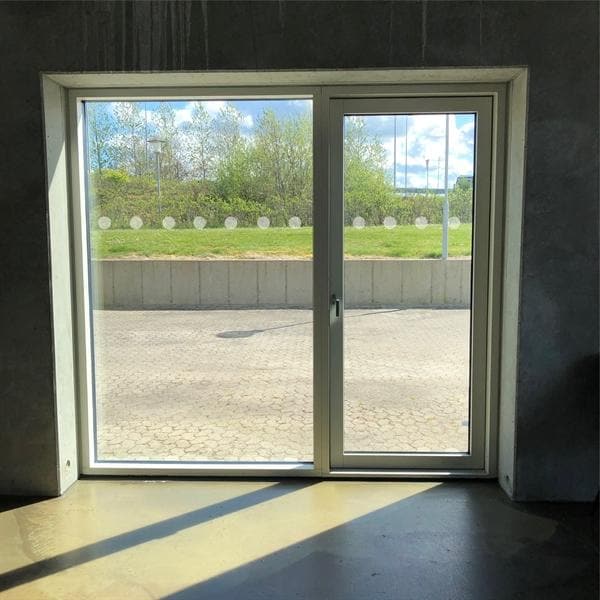 Glasdør med prikker af markeringsfolie, udsigt til flisebelagt område og grønne træer.